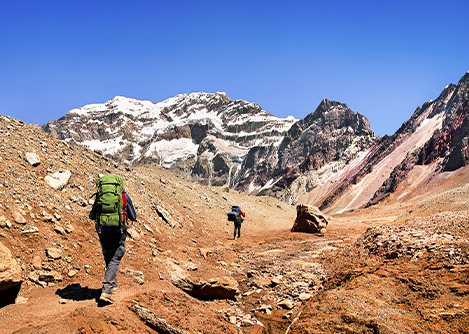 Hikers walking through a reddish and rocky landscape with mountains visible in the distance.