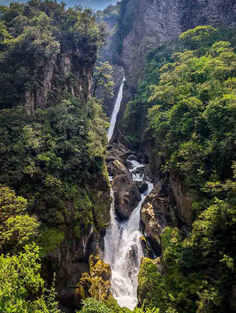 A waterfall, Pailón del Diablo, falls down a rocky cliff surrounded by lush vegetation. 