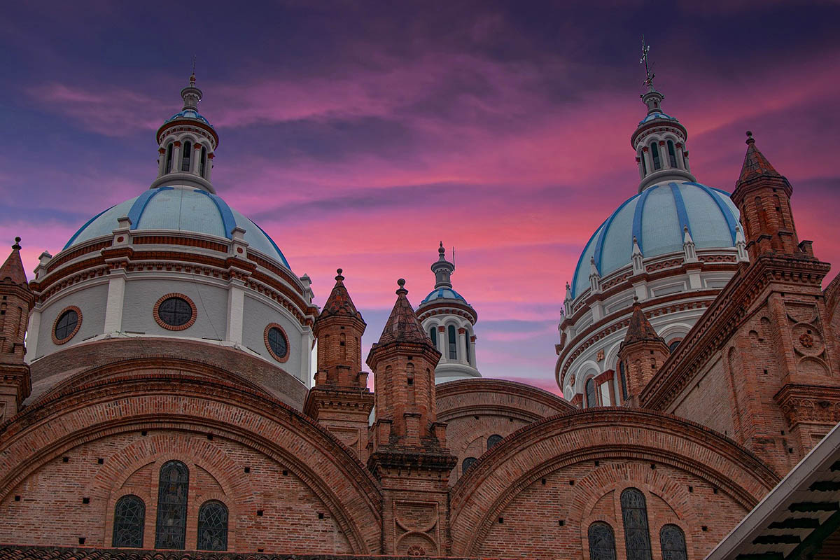 A brown church with blue domes on top and a pinkish-purple sunset behind in Cuenca, Ecuador.