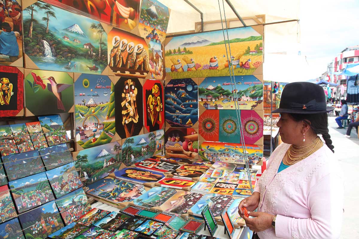 A woman wearing a bowler hat sells small, colorful paintings at a market stall in Otavalo Market.