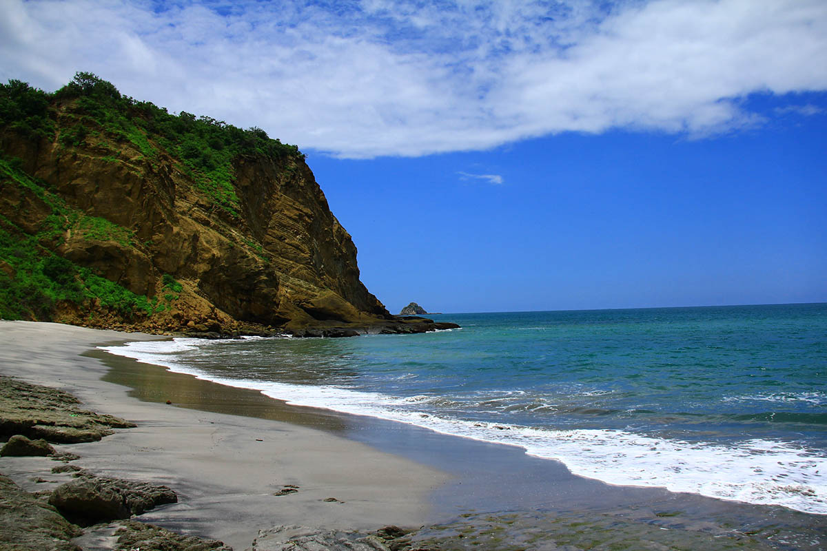 A sandy beach with rocky cliffs beyond the sandy area. Bright blue ocean and skies.