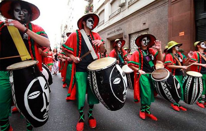 People in red, black, and green attire drumming on black and white drums in a Carnaval parade.