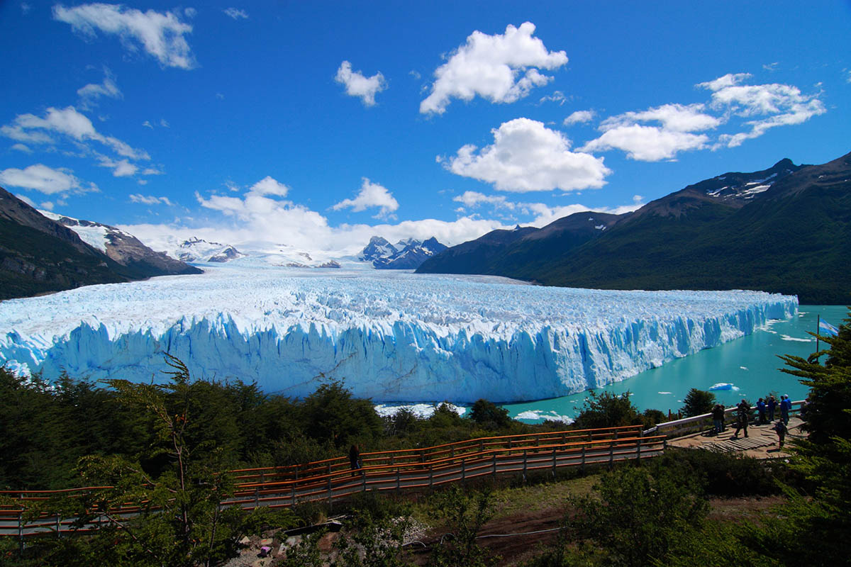 A glacier juts out in a lake surrounded by mountains in the distance.