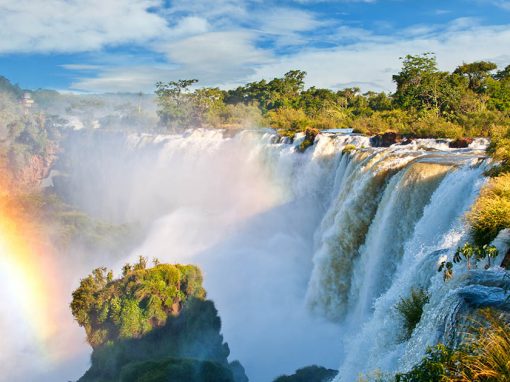 Waterfalls with a rainbow in front and and lush green trees behind.