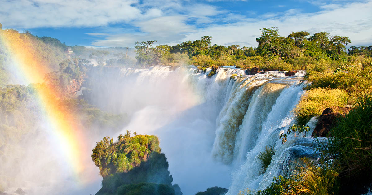 Waterfalls with a rainbow in front and and lush green trees behind.