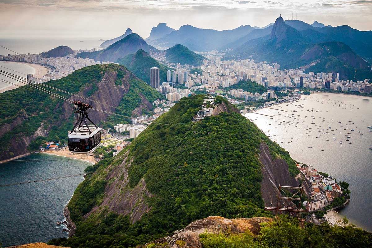 Cable cars in Rio de Janeiro pass through a green mountainous landscape along the ocean.