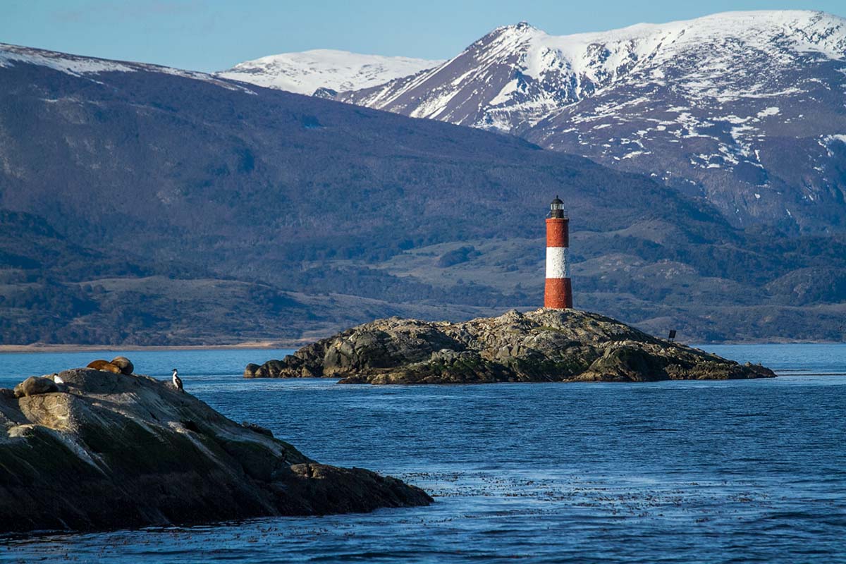 An islet with a red and white lighthouse in the middle of a bay with mountains behind in Ushuaia.
