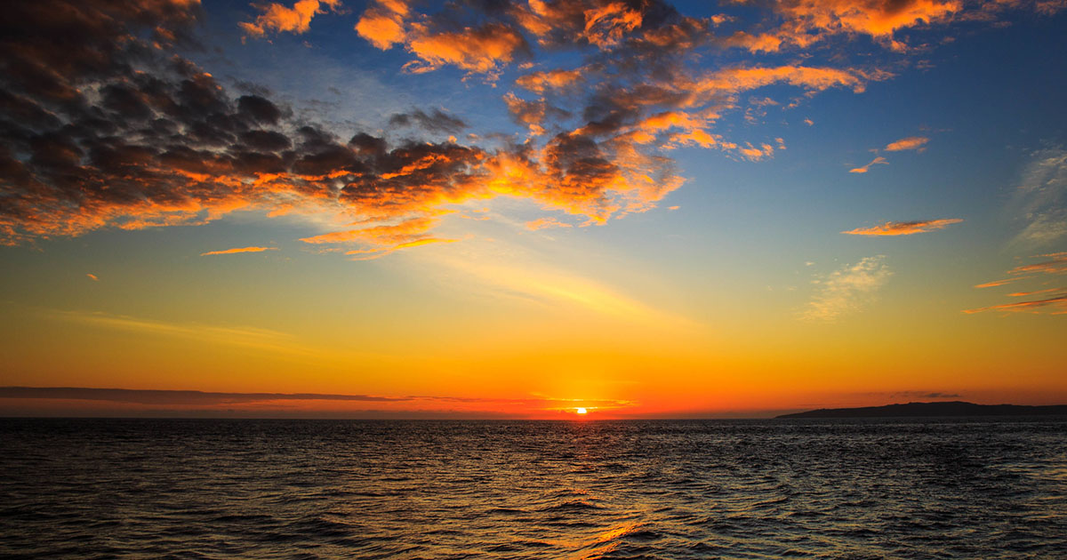 The sun sets over the ocean in the Galapagos, filling the blue sky with orange and red clouds.