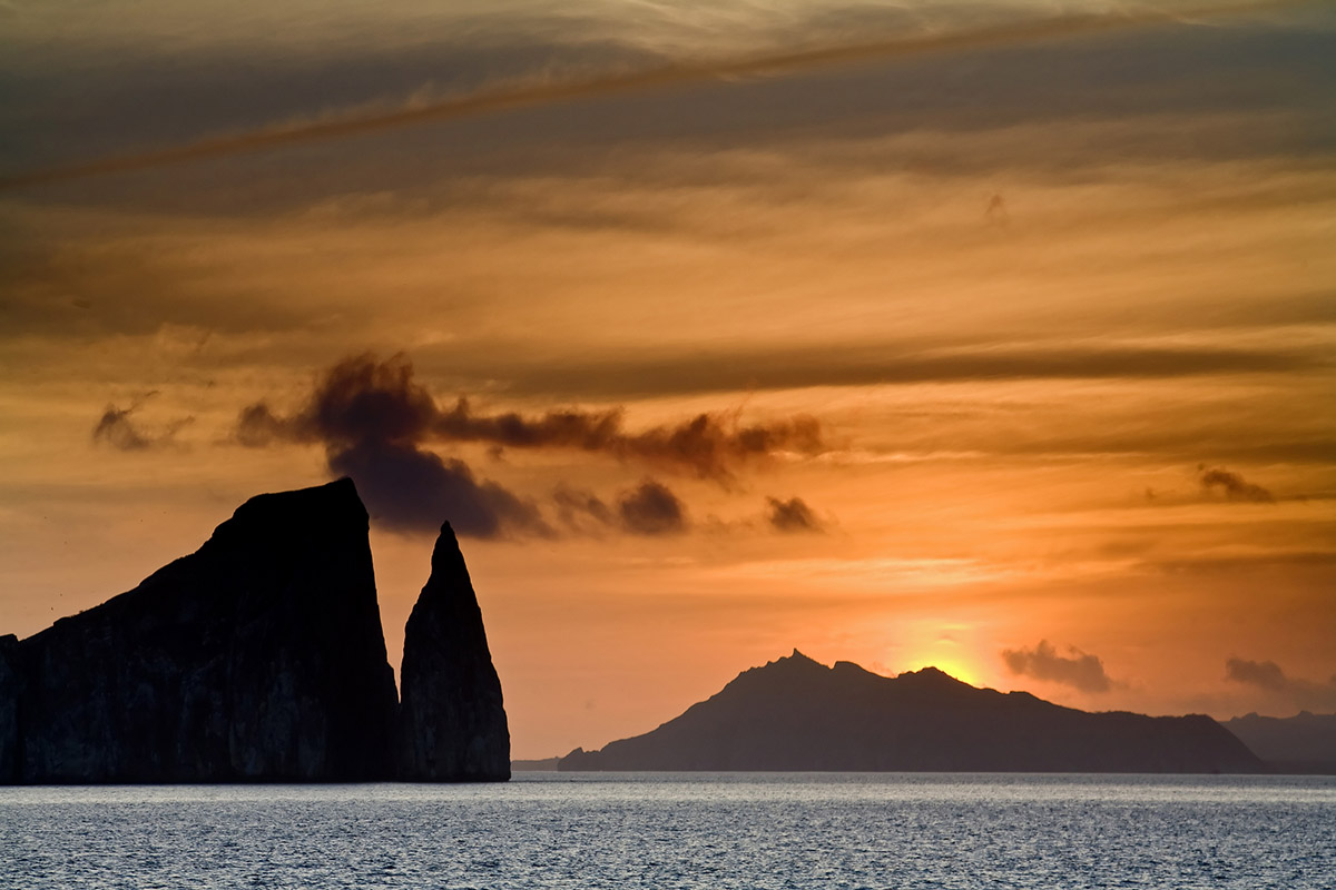 A yellow-orange sunset silhouettes Kicker Rock, a jagged rock formation jutting out of the ocean.