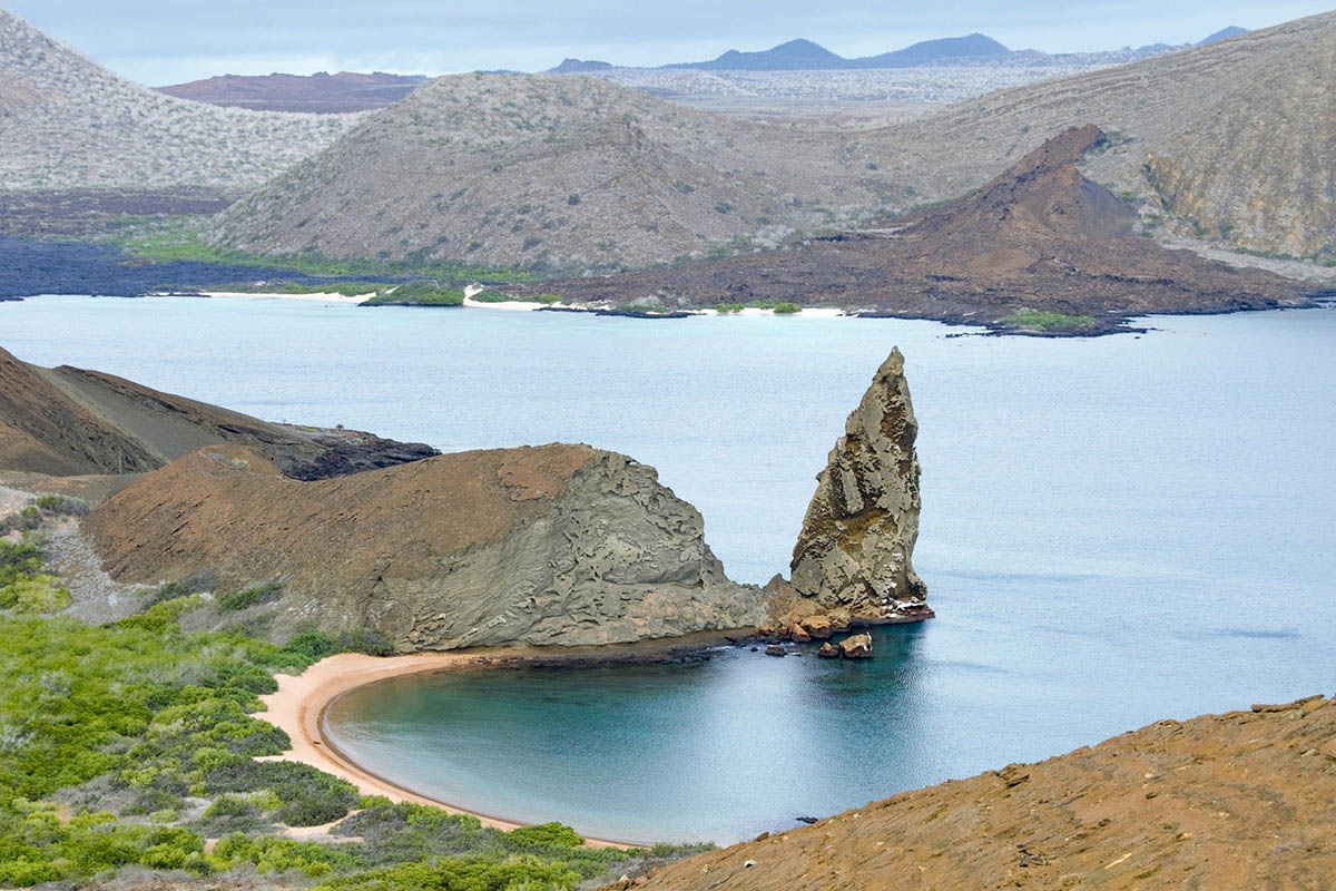 A giant sharp rock juts out from a bay on Bartolome Island. Mountains can be seen in the distance.