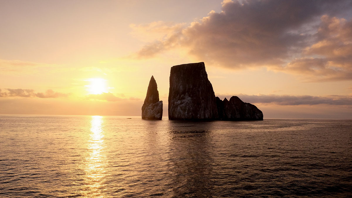 A sunset is reflected on the ocean, two large rocks jut out of the water.