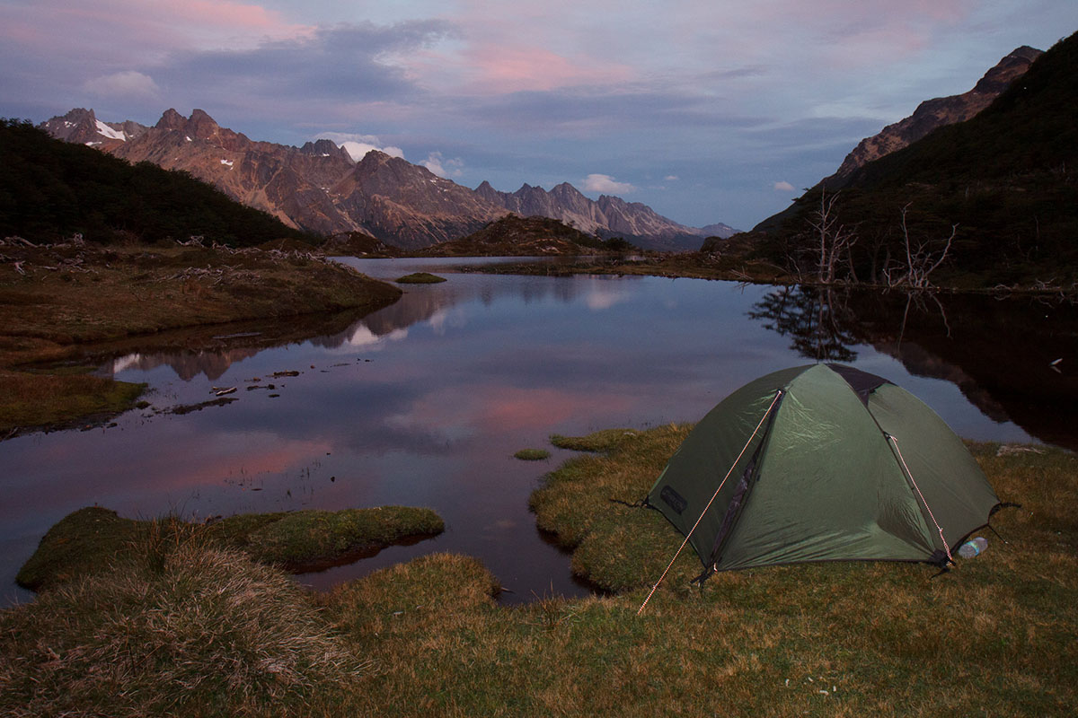 A green tent is pitched on the edge of a lake with mountains in the distance.