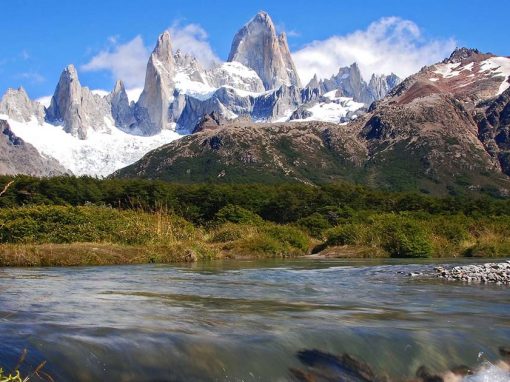 Three stone peaks surrounded by snow tower over foothills and a river.