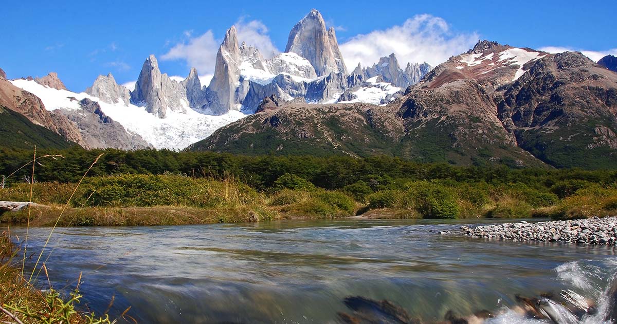 Three stone peaks surrounded by snow tower over foothills and a river.