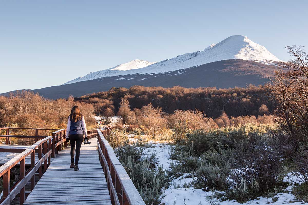 A woman with a camera walks on a wooden pathway in Tierra del Fuego National Park’s snowy scenery.