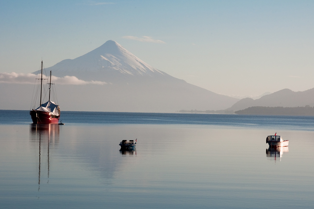 A sailboat and two dinghies sit in a calm harbor with a snowcapped volcano in the distance.