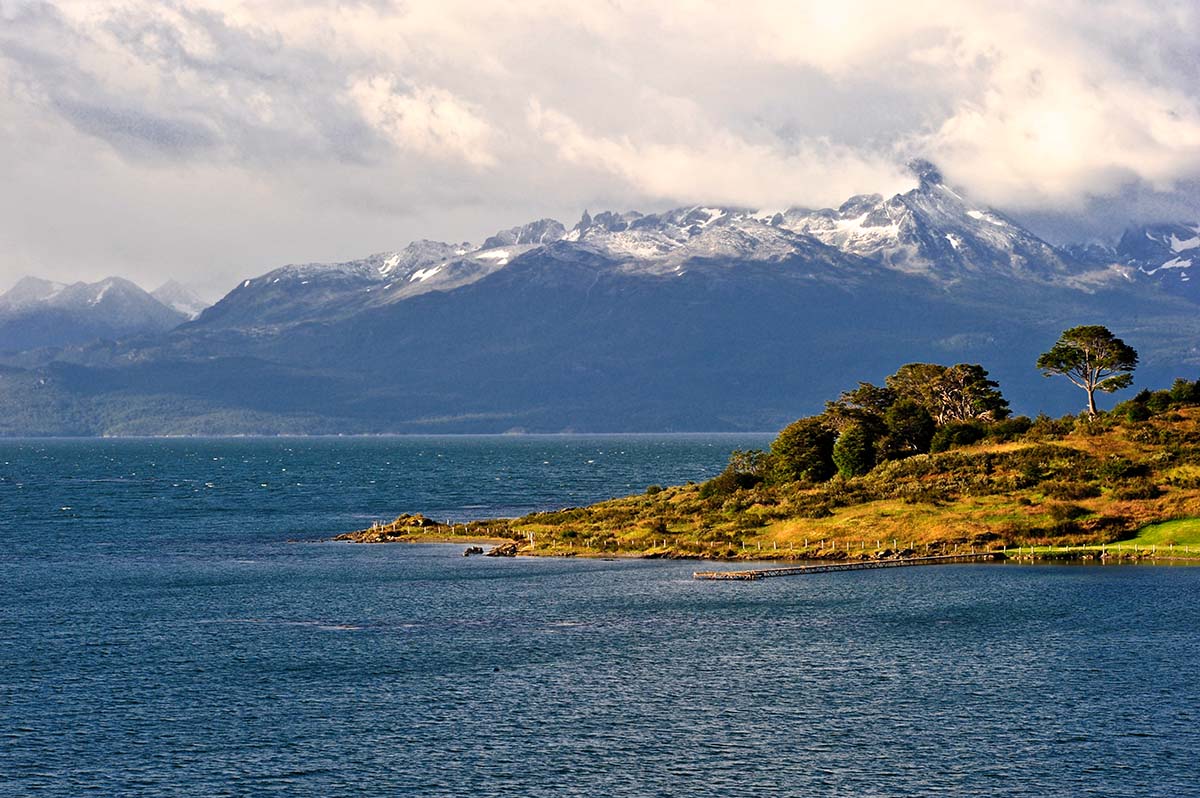 A grassy hills sticks out into the sea with snowcapped mountains in the distance.