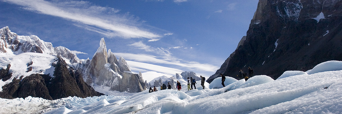 A group of hikers trudge through the snow surrounded by bare granite peaks.
