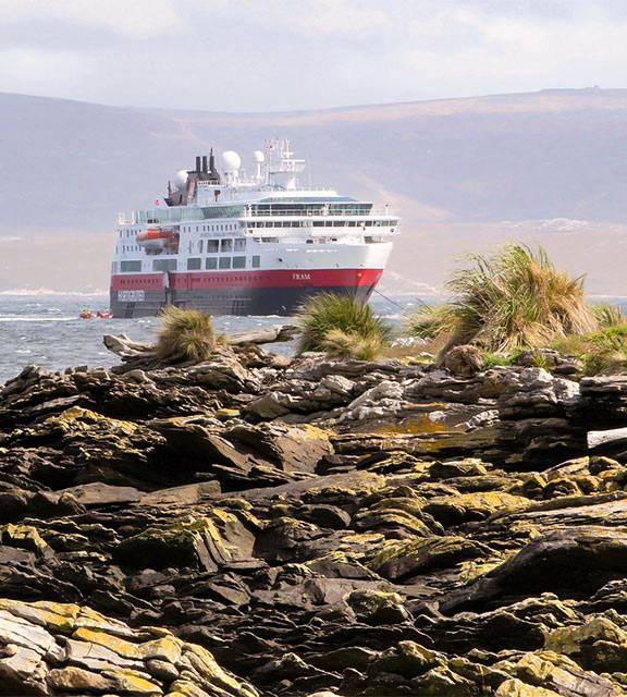 A red, white and black cruise ship approaching a rocky shore in the Falkland Islands.