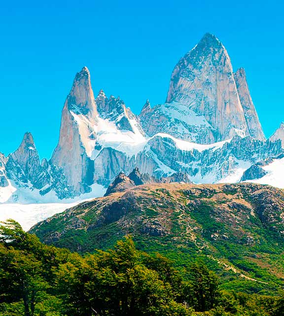 The jagged and snowy peaks of Mount Fitz Roy overlooking a hilly area covered in greenery.