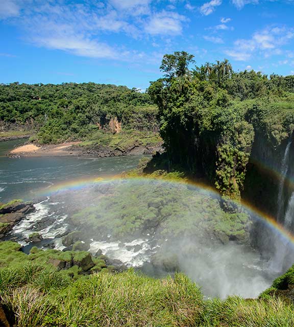 A full rainbow arch next to a waterfall at Iguazu Falls with lush greenery surrounding on all sides.