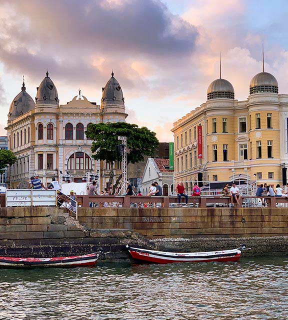 Historic buildings overlooking a river where two small row boats are docked in Recife.