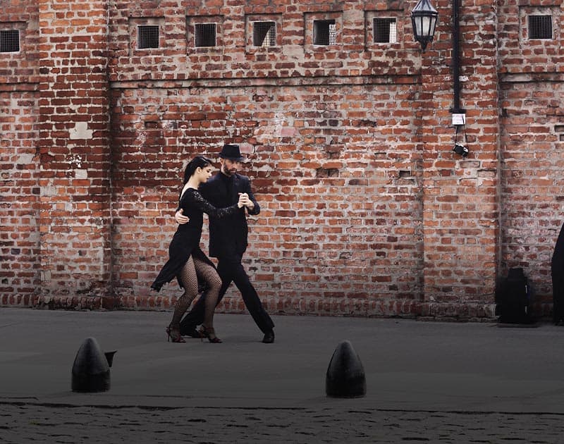 A dressed-up couple performing the tango on the street in front of a brick wall.