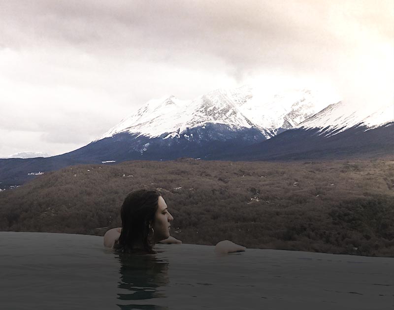A woman relaxing in an outdoor pool surrounded by a landscape of snow-capped mountains.