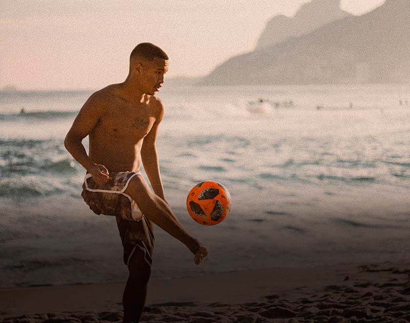 A Brazilian man kicking a soccer ball into the air with his right foot on a sandy beach in Brazil.