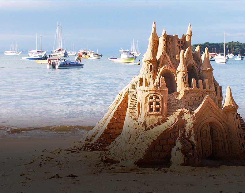 An elaborate sand castle on the beach in Buzios, with several boats anchored in the water offshore.