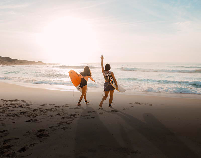 Two women carrying their surfboards towards the ocean on a sandy beach in Brazil.