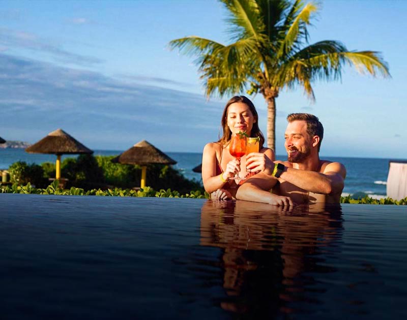 A couple enjoying some cocktails in a pool with the ocean and a palm tree in the background.