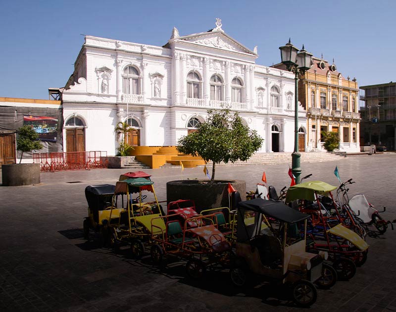 A group of pedal-powered cars overlooked by historic buildings in Iquique’s Arturo Prat Square.