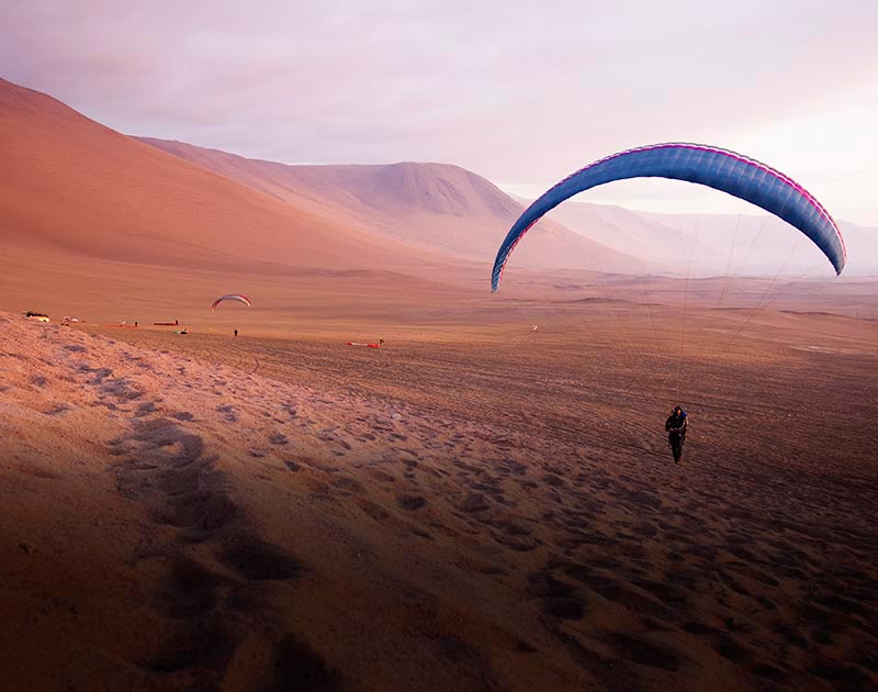 A paraglider landing on a sandy beach surrounded by desert hills near the city of Iquique.