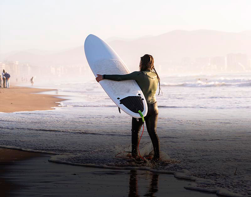 A woman wearing a wetsuit and carrying a surfboard along the beach in La Serena.