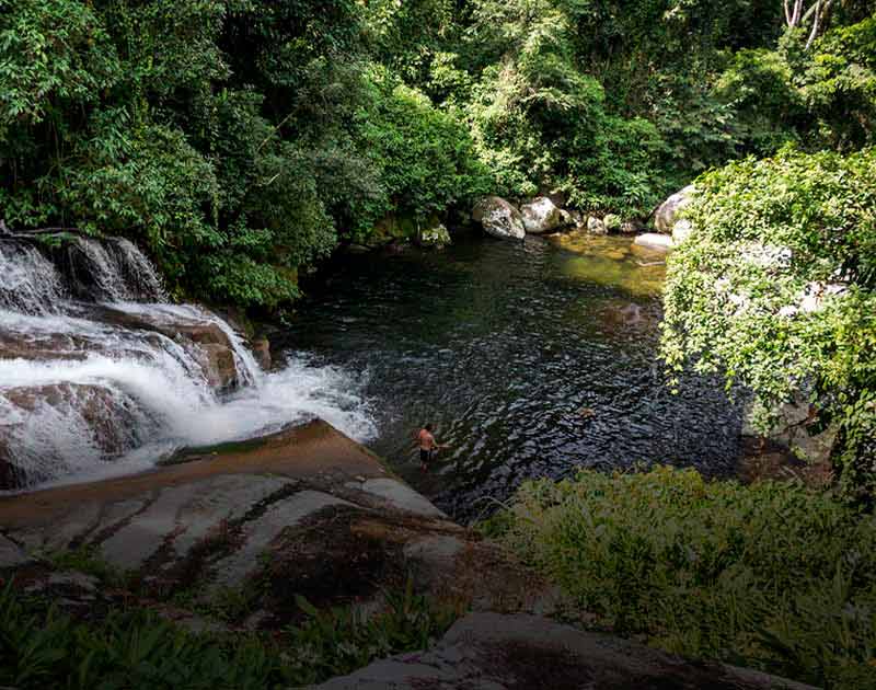 A man standing in the water beneath Pedra Branca Waterfall, surrounded by lush rainforest.