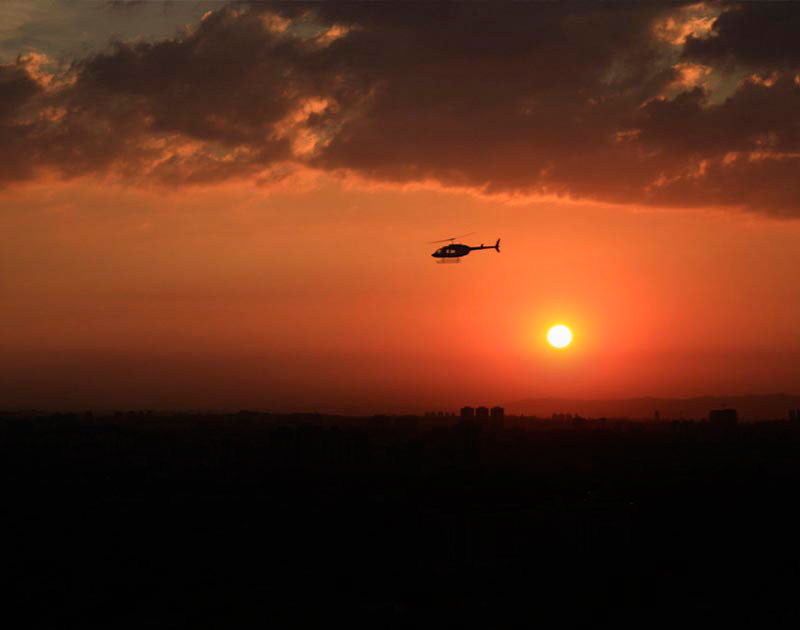 A helicopter flying over the Sao Paulo skyline, with a bright red sunset as a backdrop.