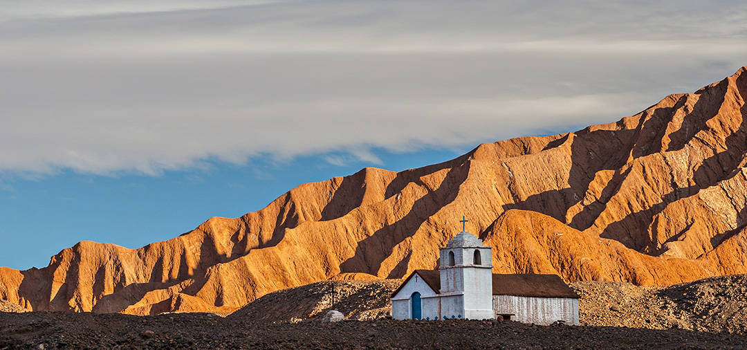 A historic church overlooked by imposing golden-colored mountains in the Atacama Desert.