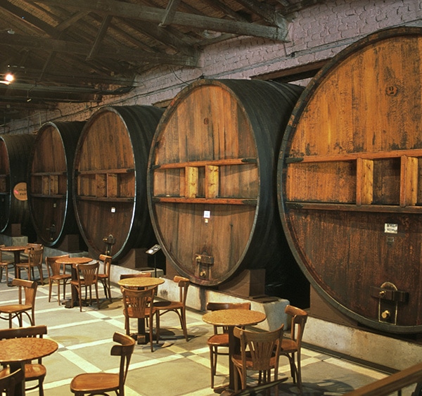 Small tables overlooked by massive wine barrels at a tasting room in a wine cellar.