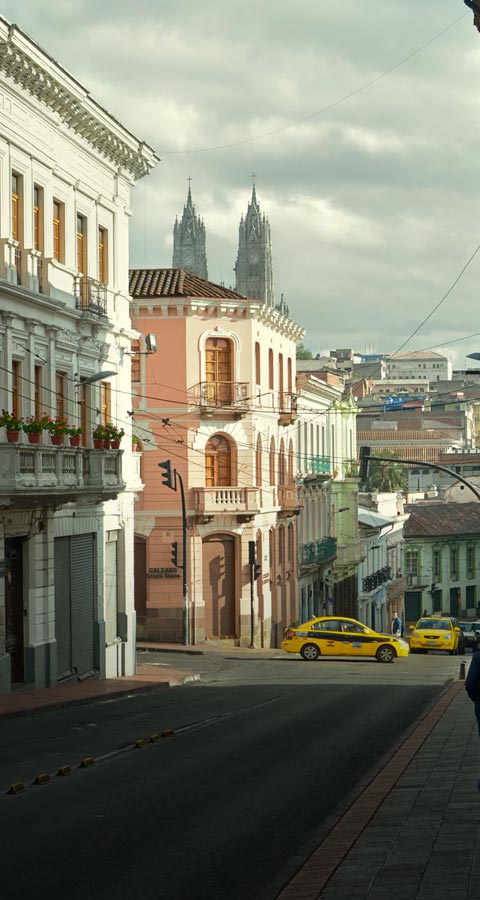 A city street in Quito lined with colorful historical buildings and cathedral towers visible in the backdrop.