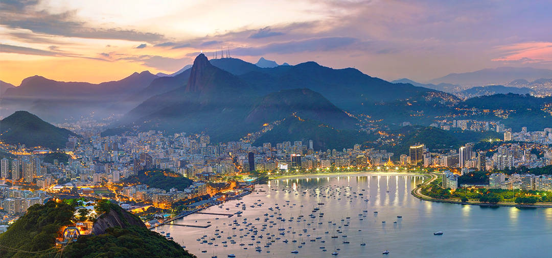 A panoramic view of Rio de Janeiro and the surrounding mountains, lit up in the evening.