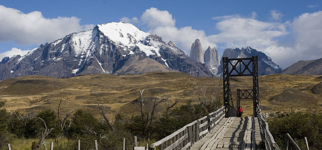 A visitor crossing a bridge in Torres del Paine National Park, with a couple of the peaks visible.