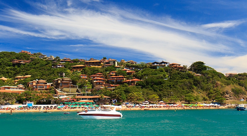 A catamaran in the water off the shore of Búzios, with a beach and many houses in the hills visible.