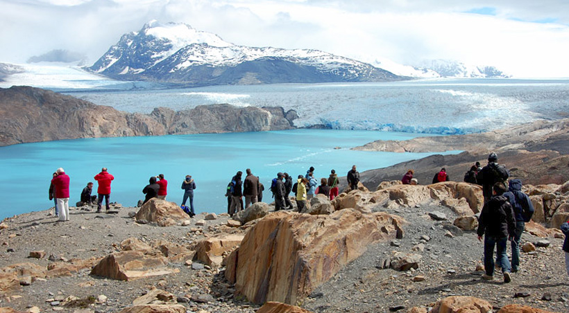 Visitors looking out at a bright blue lagoon with a glacier and snow-capped mountains behind it.