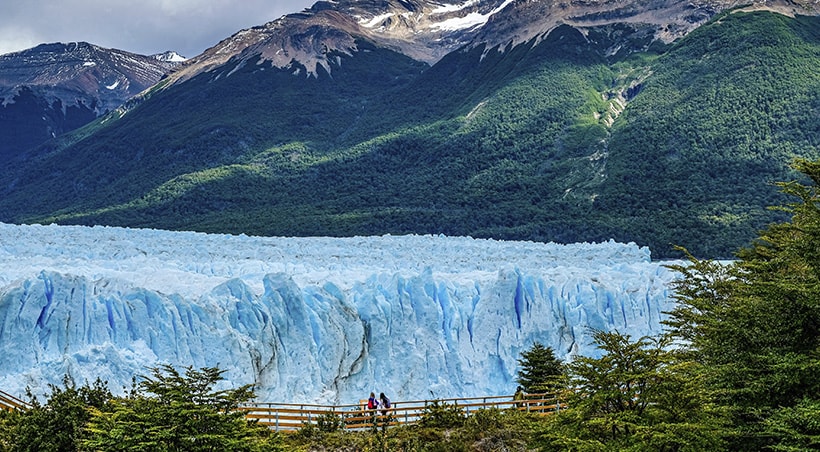 Visitors on a pathway look at a massive blue glacier, with forest-covered mountains behind.