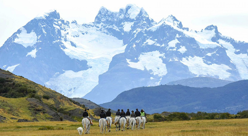 A group of visitors on horseback riding through a valley with imposing mountains towering overhead.