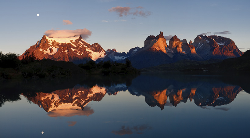 Snow-capped mountains and the back of the iconic peaks of Torres del Paine reflected in a lake.