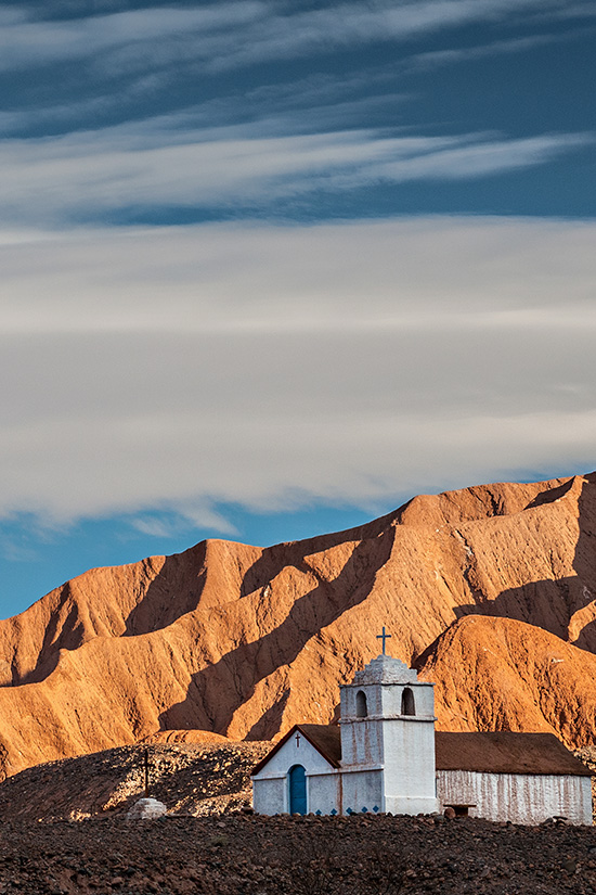 A historic church overlooked by imposing golden-colored mountains in the Atacama Desert.