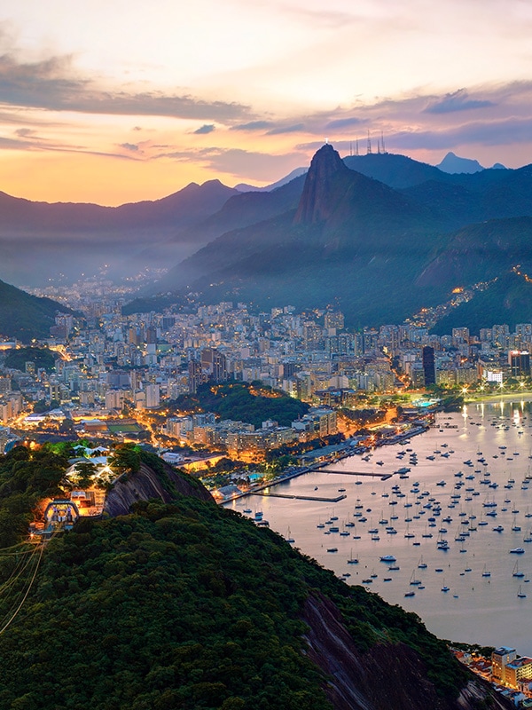 A panoramic view of Rio de Janeiro and the surrounding mountains, lit up in the evening.
