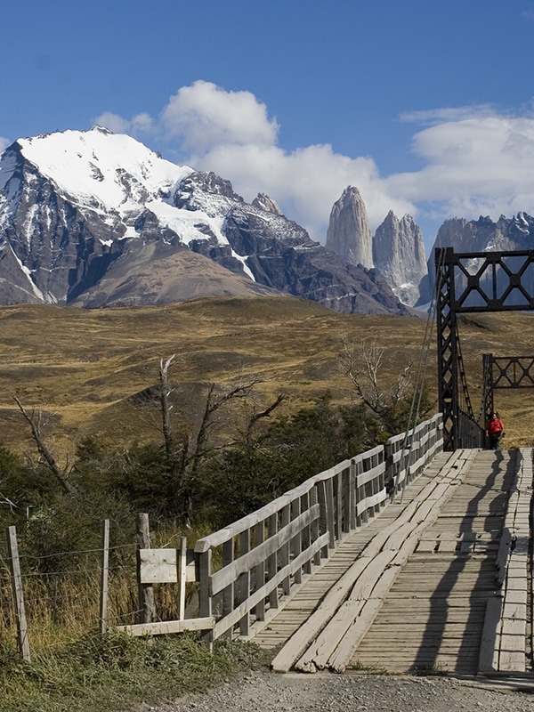 A visitor crossing a bridge in Torres del Paine National Park, with a couple of the peaks visible
